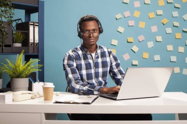 Erkek yardım hattı operatörü masada oturuyor, kameraya bakıyor, uzun çalışma saatlerinden dolayı yorgunlukla boğuşuyor. Eğri gözlükleri ve kulaklığı olan siyahi bir adamın portresi, fazla mesai yükünden boşalmış gibi görünüyor.
