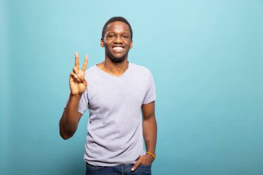Genç siyah erkek model bir eli cebinde poz verirken diğeriyle barış işareti yapıyor. Gündelik giysiler içinde Afro-Amerikalı bir adam, iki parmağıyla V işareti yapıyor ve kameraya gülümsüyor..