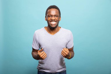 Neşeli genç adam yumruğunu kaldırıyor, kendini güçlü ve kendinden emin hissediyor, başarıyı ve gücü simgeliyor. Heyecanlı Afrikalı Amerikalı adam elleriyle tezahürat yapıyor, başarılarını kutluyor..