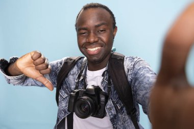 Erkek fotoğrafçının pov 'u izole edilmiş arka plana şakayla başparmak kaldırıyor. DSR kamerası ve sırt çantasıyla Afrikalı Amerikalı vlogger, anlaşmazlığı işaret ederken kendini kaydediyor..