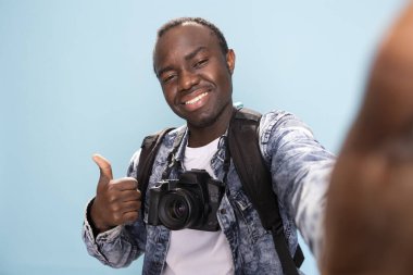 Genç siyahi bir adam pov fotoğrafı çekiyor, baş parmağıyla bir anlaşma hareketi yapıyor ve parlak bir şekilde gülümsüyor. Boynunda kamera olan Afrikalı Amerikalı fotoğrafçı mavi arka plandaki stüdyoda selfie çekiyor..