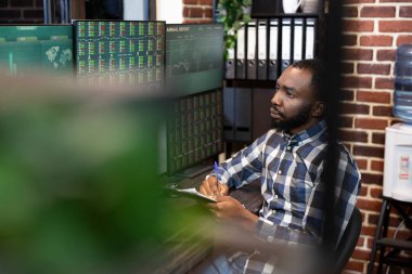 Erkek Forrex tüccarı masada oturmuş, canlı ticaret oturumunu gösteren ekranlara bakıyor ve tuğla duvar ofisinde notlar alıyor. Kalem ve pano ile ilgili finans uzmanı, hisse büyümesi istatistiklerini gözden geçiriyor.