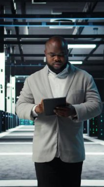 Veritabanı kontrol listesindeki tabletlere bakan dikey video uzmanı. Afrika kökenli Amerikalı bir adam sunucu çiftliğinde yürüyor, cihaz ve kamera A 'nın belgelerini okuyor.