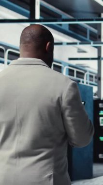 Sunucu çiftliğinde yürüyen dikey video çalışanı cihaz ekranındaki ekipman ölçümlerini analiz ediyor. Afro-Amerikan veri merkezi mühendisi, altyapı donanımları tarafından toplanan verileri kullanarak tablet üzerinde analiz yapıyor.