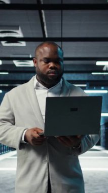 Dikey video mühendisi sunucu odasında yürüyor dizüstü bilgisayardaki yazılımı kullanarak depolama sıralarını yedekliyor. Afro-Amerikan veri merkezi mühendisi aygıt kullanarak hataları düzeltmek için donanım kayıtlarını tarıyor