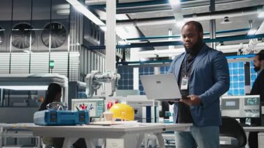 Siyah güneş paneli mühendisi yüksek teknolojili bir tesiste teftiş yapıyor. Laptoptaki verimlilik ve üretim çizgisi ölçülerine bakıyor. Sürdürülebilirlik operasyonları konusunda uzman. Kamera B.