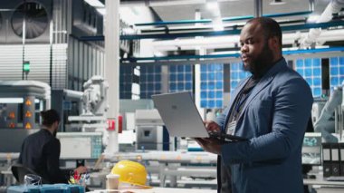 Bir güneş paneli tesisinde Afrika kökenli Amerikalı yönetici performans ve verimlilik ölçümlerini izliyor, gerçek zamanlı kalite kontrolü ve stratejik iş geliştirme ile liderliği birleştiriyor. Kamera B.