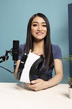 Kadın teknoloji blogcusu masada oturmuş, kameradaki VR kulaklığı inceliyor. Ürünü elinde tutarken mikrofona konuşuyor, profesyonel bir stüdyoda onun özelliklerini gösteriyor..