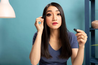 Asyalı makyaj sanatçısı çevrimiçi izleyiciler için makyaj dersi çekerken kozmetik ürün uyguluyor. Kadın içerik yaratıcısı masasında oturmuş canlı yayın sırasında rimel uygulaması gösteriyor..