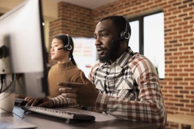 Dinlenmiş kulaklık takan Afrikalı Amerikalı bir adam bilgisayar ekranındaki bilgileri incelerken müşteri desteği sağlıyor. Siyah erkek çalışan mikrofona konuşuyor, çevrimiçi müşteriye yardım ediyor..