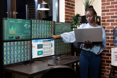 Afro-Amerikalı borsacı masanın başında dizüstü bilgisayardaki ön-pazar verilerini ve profesyonel gece ticaret ortamındaki birkaç monitörü karşılaştırıyor. Teknolojiden anlayan bir ofiste çalışan siyahi kadın..