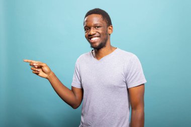 Neşeli genç adam sol tarafını işaret ediyor, mavi bir arka plana karşı duruyor. İşaret parmağıyla hareket eden gülümseyen erkek bir reklamda promosyon yapıyor. Reklam kavramı.