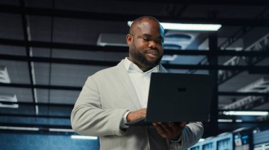 Sunucu odasında yürüyen bir mühendis, dizüstü bilgisayardaki yazılımı kullanarak depolama sıralarını yedekliyor. Afro-amerikan veri merkezi mühendisi aygıt, kamera B kullanarak hataları düzeltmek için donanım kayıtlarını tarıyor