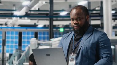 Siyah güneş enerjisi uzmanı dizüstü bilgisayardaki verileri inceliyor, üretim tesisinin performansını inceliyor ve yeniliği sembolize ediyor. Sanayi uzmanı üretim hattını denetliyor. Kamera A.