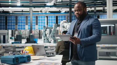 Bir güneş enerjisi üretim tesisinde çalışan Afrikalı Amerikalı teknisyen şirket denetimi ve ekipman üretimini yansıtmak için takım elbise giymiş operasyonları değerlendiriyor. Temiz enerji. Kamera B.