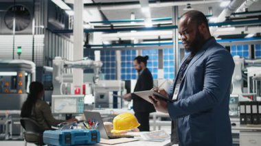 Siyah usta mühendis bir fabrikada fotovoltaik üretim hattını denetleyerek, kurumsal hedefler ve alternatif enerji standartlarını karşılamak için başarılı bir üretim süreci sağladı. Kamera B.