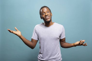 Genç siyahi bir adam stüdyoda jest yapıyor, omuzlarını kaldırıyor ve kararsızlığını dile getiriyor. Afrikalı Amerikalı adam kolları açık duruyor, izole edilmiş bir geçmişe karşı şaşkınlık içinde omuz silkiyor..