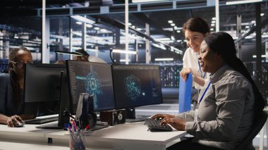 Veri merkezi ekip lideri ve bilişim uzmanı yapay zeka ekipmanlarının performans gösterge panellerini inceliyor. Sunucu odası yöneticisi sistem yöneticisiyle ilgilenir. Ekranda inceleme makinesi öğrenme algoritmaları, kamera A