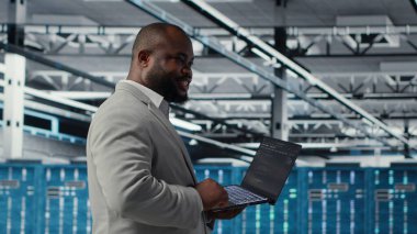 Cheerful data center system administrator reviews code for automation scripts. Jolly server room employee running diagnostics on equipment, doing predictive error detection, camera B