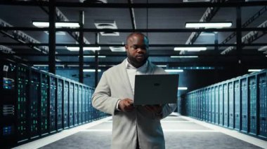 Sunucu odasında yürüyen bir mühendis, dizüstü bilgisayardaki yazılımı kullanarak depolama sıralarını yedekliyor. Afro-Amerikan veri merkezi mühendisi cihaz, kamera A kullanarak hataları düzeltmek için donanım kayıtlarını tarıyor.