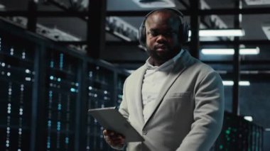 Sunucu odası programcısı kulaklık takıyor. Tabletteki yazılımı kullanarak sorun çıkarma hatası. IT uzmanı müzik dinliyor, veri merkezinde yürüyor, aygıt kullanarak hataları düzeltiyor, kamera B