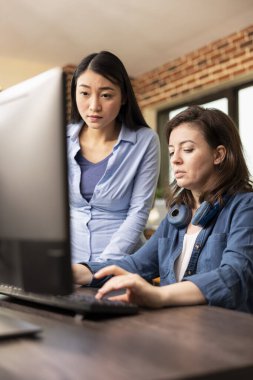 Asyalı yönetici ayakta dikilirken ve masaüstü monitöründeki çalışmaları incelerken, beyaz çalışanın klavyede daktilo yazması. Bilgisayar kullanan iki profesyonel kadın modern ofiste yaratıcı bir tartışma yapıyorlar..