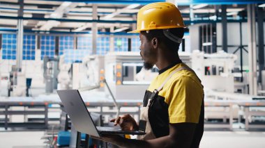 Güneş paneli tesisindeki siyah teknisyen fotovoltaik sistem performansını dizüstü bilgisayardan inceliyor. Endüstriyel santraldeki verimliliği optimize etmek için üretim hattı ölçümlerini analiz ediyorum. Kamera B.