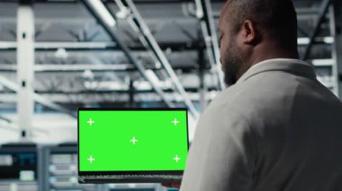 Veri merkezi programcısı yeşil ekran bilgisayarı üzerinde çalışıyor, depolama altyapısını güncelliyor. Bilgi işlem uzmanı sunucu merkezinde ağ teknolojisi ve kamera B ile dolu bir dizüstü bilgisayar kullanıyor.