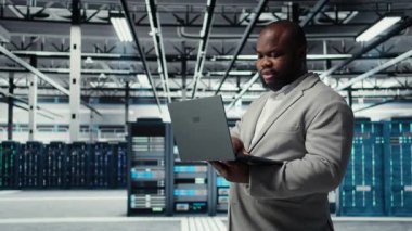 Computer scientist using laptop in data center, managing server virtualization. African american man using notebook in server room, ensuring infrastructure reliability, camera A