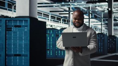 Veri merkezindeki gülümseyen mühendis dizüstü bilgisayar kullanarak sistem istikrarı için yazılım güncellemelerini yönetiyor. Sunucu çiftliğindeki mutlu çalışan performansı optimize ediyor, altyapıyı dizüstü bilgisayarla yapılandırıyor, kamera A