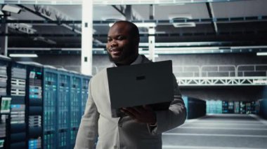 Veri merkezinde yürüyen teknisyen, yapay zeka iş yüklerinde en az gecikme için depolama cihazlarını yapılandırıyor. Afrikalı Amerikalı çalışan, sunucu çiftliğinde sinirsel ağlar kurmak için aygıt kullanıyor. Kamera A