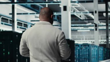 Çalışanların sunucu çiftliğinde yürümesi, cihaz ekranındaki ekipman ölçümlerini analiz etmesi. Afro-Amerikan veri merkezi mühendisi tablet üzerinde analiz yapıyor. Altyapı donanımları ve kamera B ile elde edilen verileri kullanıyor.