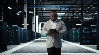 Bilgi işlem uzmanı veri merkezinin kontrol listesi evraklarını inceliyor. Afrika kökenli Amerikalı bir adam sunucu çiftliğinde yürüyor, cihaz ve kamera A 'nın belgelerini okuyor.
