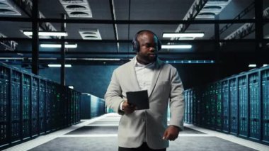 Sunucu odası programcısı kulaklık takıyor. Tabletteki yazılımı kullanarak sorun çıkarma hatası. IT uzmanı müzik dinliyor, veri merkezinde yürüyor, aygıt kullanarak hataları düzeltiyor, kamera A