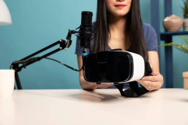 Profesyonel stüdyo ortamında, bir kadın ürün eleştirmeni bir VR kulaklığı göstermektedir. Cihazı, internetteki teknik izleyicilerine mikrofona konuşurken kameraya sunuyor..