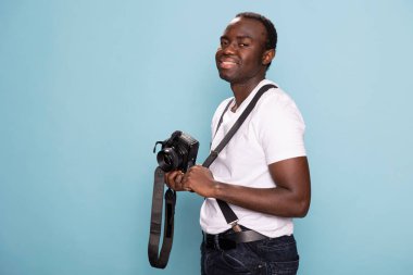 Afrikalı Amerikalı bir adam bir elinde kamera, diğer elinde de pantolon askısı ile güvenle duruyor. Sıradan giyinmiş siyah erkek modelin portresi, DSLR 'ıyla poz veriyor..