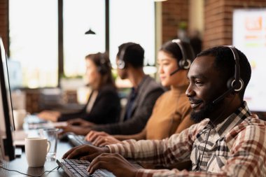 Afrika kökenli Amerikan çağrı merkezi ajanına yakın çekim. Uzak müşterilere danışmanlık yaparken bilgisayarındaki bilgileri gözden geçiriyor. Bilgisayarlarını kullanan çoklu etnik pazarlamacılarla modern çalışma alanı.