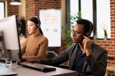 Çevrimiçi pazarlama sunumu sırasında dikkatlice dinleyen, kulaklık takan siyah erkek girişimci. Afrikalı Amerikalı tekerlekli sandalye kullanıcısı müşterileriyle duvarlarla çevrili bir ofiste iletişim kuruyor..