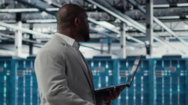 Sistem istikrarını yükseltmek için dizüstü bilgisayar kullanan gülümseyen bir veri merkezi programcısı. Neşeli Afro-Amerikan sunucu merkezi bilişim uzmanı kodlama, aygıt ile performansı optimize etme, düzenek, kamera B