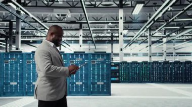 Sistem istikrarı için yazılım güncellemelerini yönetmek için tablet kullanan veri merkezindeki programcı. Sunucu odasında Afrikalı Amerikalı bir adam performansı cihazla optimize ediyor, altyapıyı yapılandırıyor, kamera A