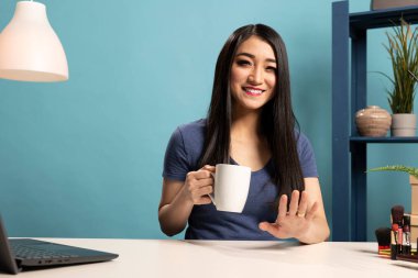 Mavi arkaplanı olan bir stüdyoda canlı yayın sırasında gülümseyen ve çevrimiçi izleyicilere konuşan güzellik uzmanı portresi. Neşeli blogcu masaya oturmuş, elinde kahve fincanı tutarak kameraya bakıyor..