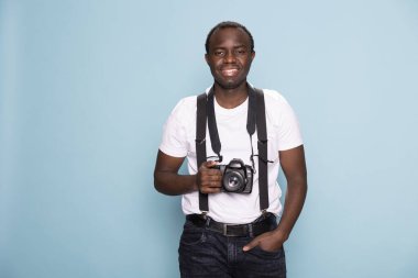 Siyah fotoğrafçı boynundaki kamerayla poz veriyor, profesyonellik yayıyor. Mavi bir arka plana karşı duruyor, gündelik modasını ve yaratıcı özünü vurguluyor..