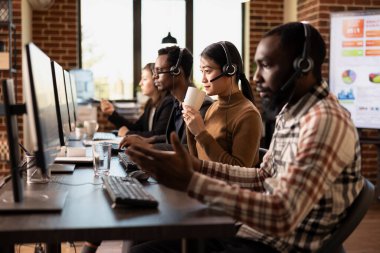 Asyalı kadın çağrı merkezi ajanı bir fincan kahve içiyor ve bilgisayarındaki müşteri bilgilerini inceliyor. İş yerinde çok ırklı pazarlamacılarla modern çalışma alanı, çevrimiçi müşterilerle konuşuyor.