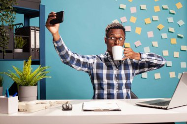 Siyah adam çalışma masasında oturmuş, kahve içiyor ve kendini akıllı telefonuyla kaydediyor, iş gününü belgeliyor. Afrikalı Amerikalı iş danışmanı selfie çekiyor ve bir fincan çayın tadını çıkarıyor..