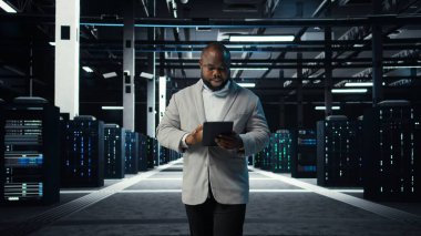 Bilgi işlem uzmanı veri merkezinin kontrol listesi evraklarını inceliyor. Afrika kökenli Amerikalı bir adam sunucu çiftliğinde yürüyor, cihaz ve kamera A 'nın belgelerini okuyor.