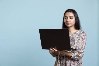 Hintli genç yetişkin e-posta yazıyor ya da stüdyoda dizüstü bilgisayarında yazı yazıyor, bilgisayardaki profesyonel görevlerini çözüyor. Günlük odaklı kişi internette mavi arkaplana karşı geziniyor.