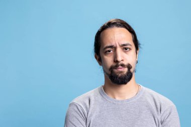 Ciddi bir insan kamerada hayal kırıklığı yaratan yüz ifadeleri yapıyor, kollarını mavi arka planda kavuşturup düşünüyor ve poz veriyor. Stüdyoda özgün bir erkek model..