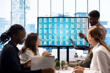 Kurul odasındaki konferans masasında işbirliği yapan iş ortakları, pazar eğilimleri ve ekonomik tahminlerin ayrıntılı bir analizini sunmak için akıllı teknoloji ve aylık istatistiklerden yararlanıyorlar..