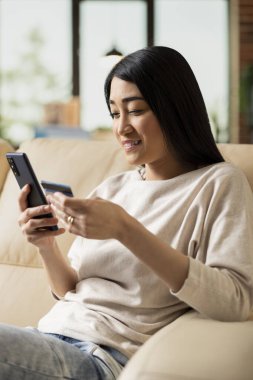 Neşeli Asyalı girişimci, internet bankacılığı için akıllı telefon ve banka kartını kullanarak kanepeye oturur. Modern ev ofisinden dijital ödemeyi gülümseyerek tamamlayan uzak bayan yöneticiye yakın çekim.