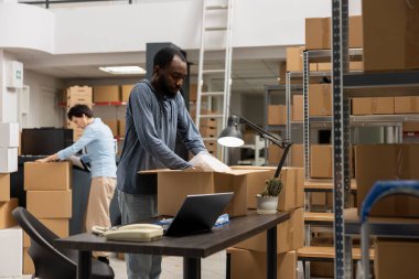 Siyah erkek yönetici, yerel bir dağıtım merkezinde çalışarak depoda lojistik siparişi vermek için dizüstü bilgisayar kullanıyor. Kutular ve envanter eşyalarıyla dolu malzeme odası, e ticaret..